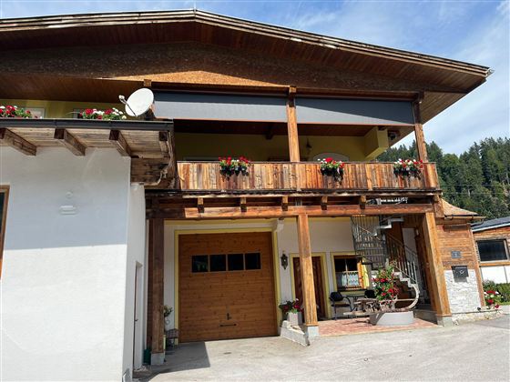 A traditional wooden house with a balcony and blooming plants. The facade is bright and welcoming, surrounded by a picturesque landscape.