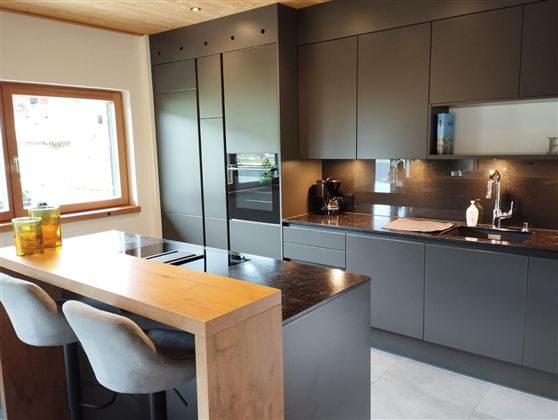 A modern kitchen with dark cabinets and an elegant countertop. At the table, there are chairs, and daylight is coming in through the window.