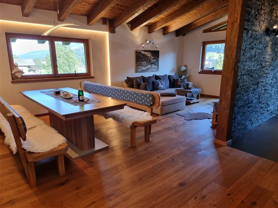 A cozy living kitchen with wooden ceilings and a large dining area. In the background, there is an inviting sitting area with a view of the countryside.