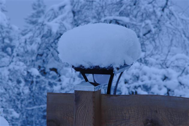 Eine Laterne mit einer dicken Schneeschicht darauf steht vor einem Holzzaun. Der Hintergrund ist mit schneebedeckten Bäumen in einer winterlichen Atmosphäre.