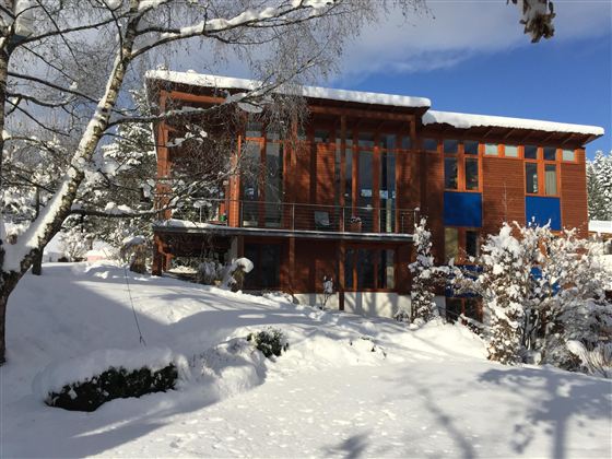 Ein modernes Holzhaus im Schnee, umgeben von winterlicher Landschaft. Der Himmel ist klar und blau, und die Bäume sind mit Schnee bedeckt.