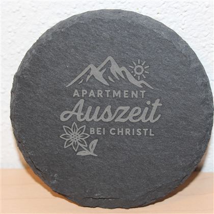 Ein runder, grauer Stein mit der Aufschrift "Apartment Auszeit bei Christl". Darüber sind stilisierte Berge und eine Sonne sowie eine Blume abgebildet.