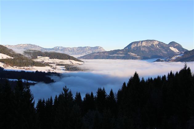 Eine beeindruckende Berglandschaft mit schneebedeckten Gipfeln. Dichte Nebel bedeckt das Tal, während der klare Himmel über der Szenerie strahlt.