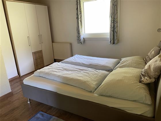 Ein helles Schlafzimmer mit einem Doppelbett und einem großen Fenster. Der Raum hat einen schlichten Stil mit Holzfußboden und einem Kleiderschrank.