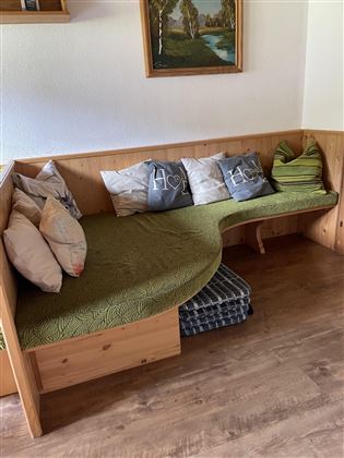 Eine gemütliche Sitzecke mit gepolsterten Kissen und einer grünen Decke. Im Hintergrund hängt ein gemaltes Bild und der Boden ist aus Holz.