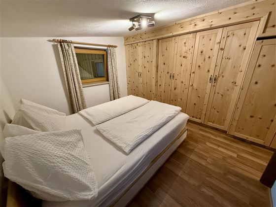 Ein gemütliches Schlafzimmer mit einem Doppelbett und Holzschränken. Große Fenster sorgen für Licht und eine helle Atmosphäre.