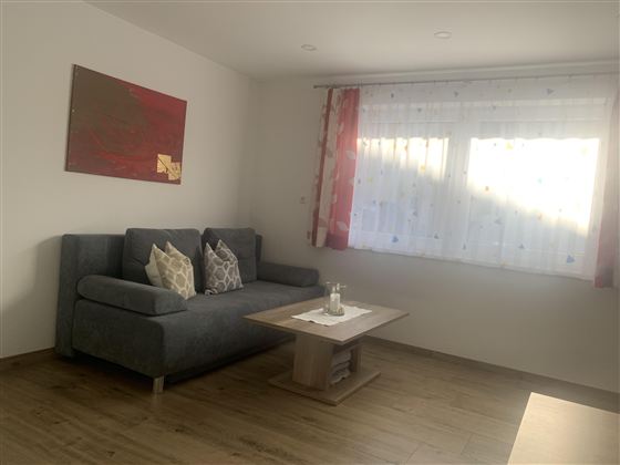 Ein gemütliches Wohnzimmer mit einem grauen Sofa und einem Tisch. Die Fenster sind hell und mit durchsichtigen Gardinen dekoriert.