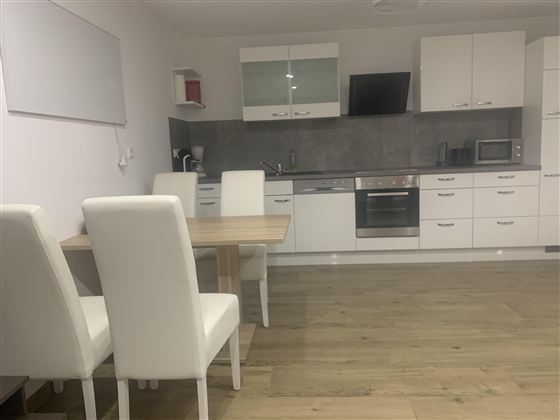 Eine moderne Küche mit weißen Möbeln und Holzfußboden. Am Tisch stehen vier weiße Stühle und die Kücheneinrichtung ist schlicht und funktional.