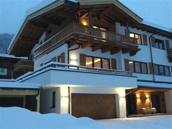 Ein modernes Chalet im Schnee, umgeben von einer winterlichen Landschaft. Die Fassade ist hell und strukturiert, mit einem großen Balkon und warmem Licht.