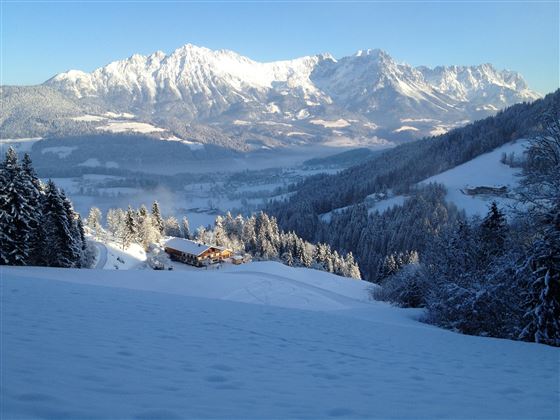 Een winterlandschap met besneeuwde bergen en een rustig dal. Op de voorgrond is een charmant houten huis te zien.