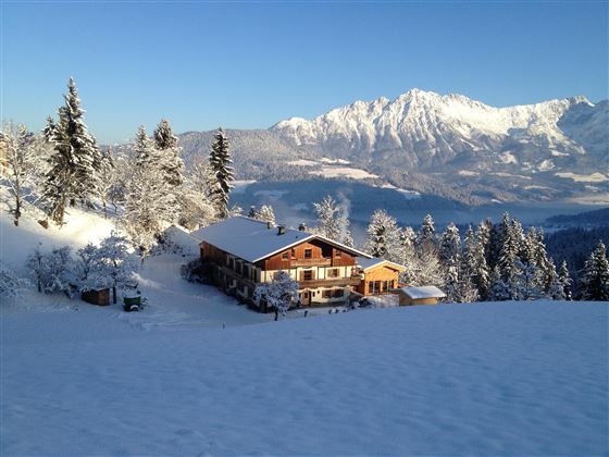 Een prachtig, sneeuwbedekt landschap met een gezellig huis. Op de achtergrond zijn majestueuze bergen en een heldere, blauwe lucht te zien.