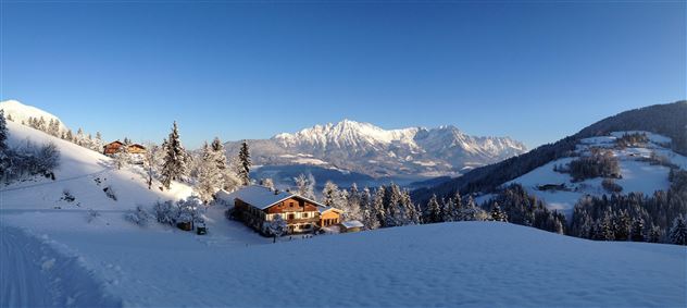 Een winterlandschap met met sneeuw bedekte heuvels en een heldere blauwe lucht. Op de voorgrond staan charmante houten huizen en besneeuwde bomen.