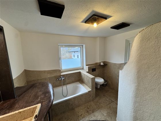 Ein modernes Badezimmer mit einer Badewanne, einem WC und einem Fenster. Die Wände sind in neutralen Farben gehalten, und der Boden ist mit Fliesen ausgelegt.