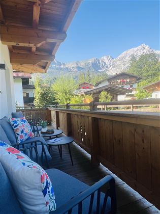 Een balkon met comfortabele zitplaatsen en kleurrijke kussens. Op de achtergrond zijn bergen en groene bomen te zien.