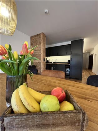 Stůl se dřevěnou krabicí plnou ovoce, včetně banánů a jablek. V pozadí je moderní kuchyň a kytice s tulipány.