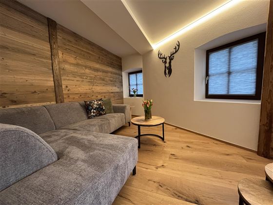 Moderní obývací pokoj s šedou pohovkou a dřevěným stolem. Na zdi visí jelení parohy a velká okna přivádějí dovnitř hodně denního světla.