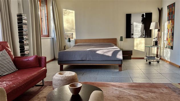 Ein modernes Schlafzimmer mit einem großen Bett und einem roten Sessel. Die Wände sind hell und es gibt einige Kunstwerke sowie einen kleinen Tisch in der Mitte.