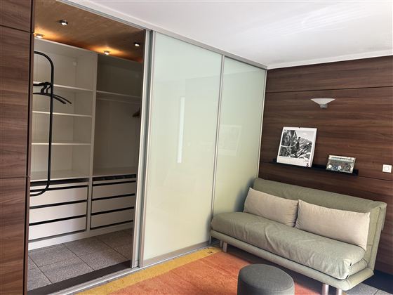 Een moderne kamer met een grijze bank en een glazen schuifdeur naar de kast. De muren zijn in een donkere houtkleur gehouden.