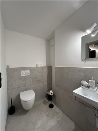 Moderní koupelna s toaletou a umyvadlem. Stěny jsou světlé a podlaha má šedé dlaždice.