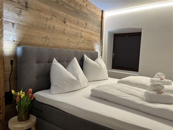 Pohodlná ložnice s velkou postelí a polštáři. Na zdi se nachází dřevěný obklad a místnost je dobře osvětlená.