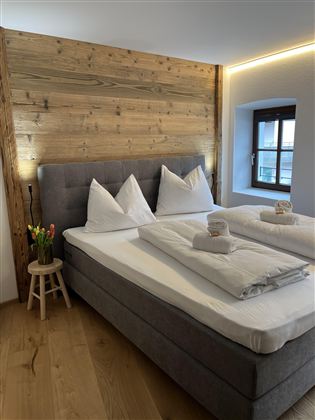 Pohodlná ložnice s manželskou postelí a dřevěným panelovým obložením. Vedle postele je malý stůl s čerstvými květinami.