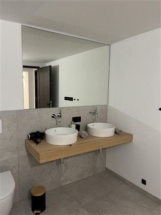 Moderní koupelna s floating dřevěnou umyvadlovou skříňkou a dvěma bílými umyvadly. Velké zrcadlo odráží světlý prostor s minimalistickým designem.