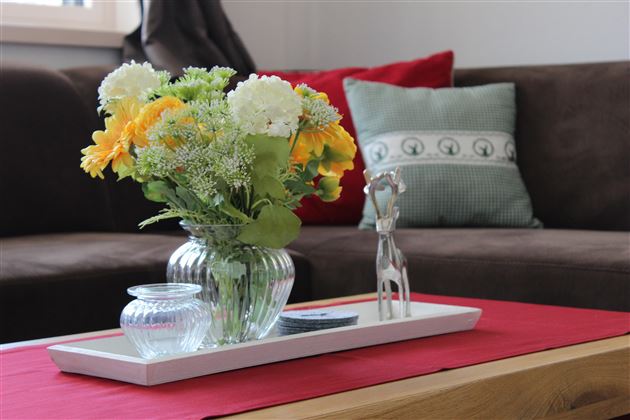 A stylishly decorated table with a flower vase and a small decorative object. In the background, a cozy sofa and cushions can be seen.