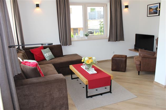 A cozy living room with a brown sofa and a red table. There is a television and plenty of daylight coming through the windows.