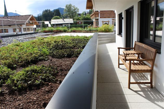 A beautiful balcony with a wooden bench and a view of a green landscape. In the background, houses and mountains can be seen.
