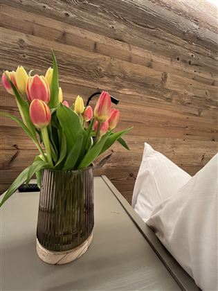 Váza s barevnými tulipány stojí na stole. V pozadí je vidět dřevěná zeď a polštář.