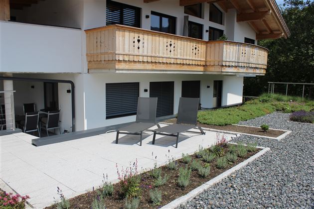 A modern house with a balcony and a terrace. On the terrace, there are reclining chairs, and they are surrounded by a beautiful garden.