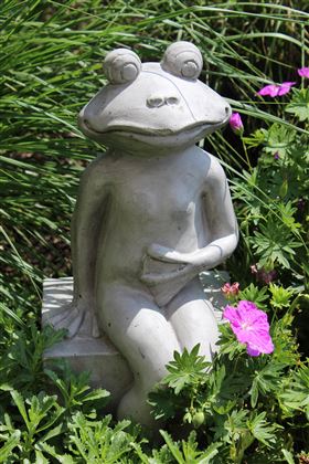 A sculpture of a seated frog made of stone, placed in a garden among green plants and colorful flowers. The frog has a relaxed body posture and a friendly expression.