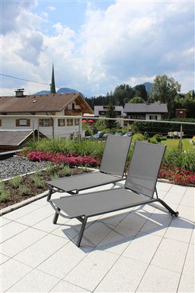 Two gray loungers on a terrace with blue sky and clouds. In the background, houses and trees are visible.