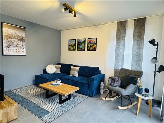 Een moderne woonkamer met een blauwe bank en een houten salontafel. De muren zijn versierd met schilderijen en er is een gezellige stoel.
