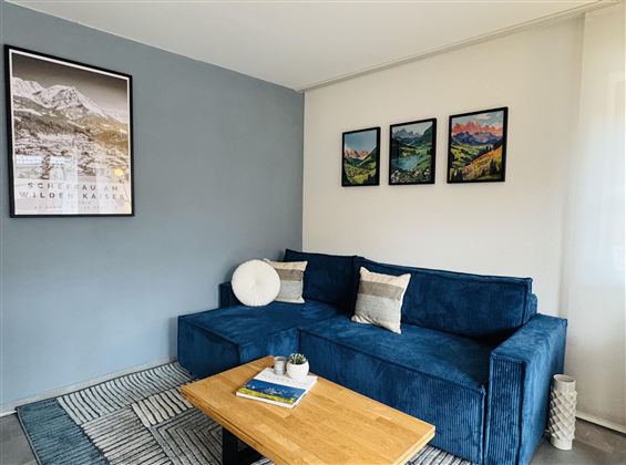 Een moderne woonkamer met een blauwe bank en een houten tafel. Aan de muur hangen foto's van landschappen en de ruimte is vriendelijk en licht ingericht.