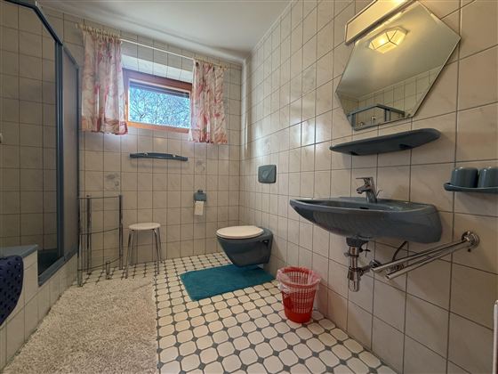 Ein Badezimmer mit gefliesten Wänden und einem großen Fenster. Es gibt eine Dusche, ein Waschbecken und eine Toilette, sowie einige Badezimmeraccessoires.