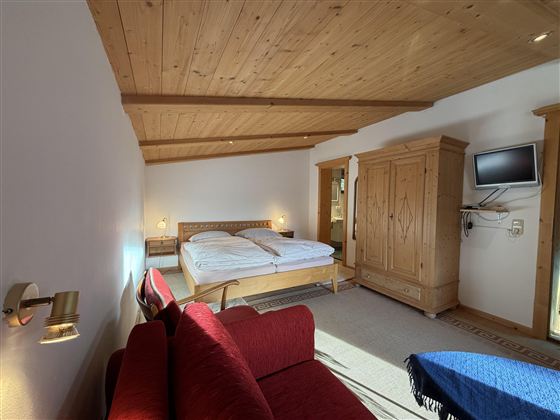 Ein helles Schlafzimmer mit Holzdecken und einem großen Bett. Es gibt eine rote Couch und einen Fernseher an der Wand.