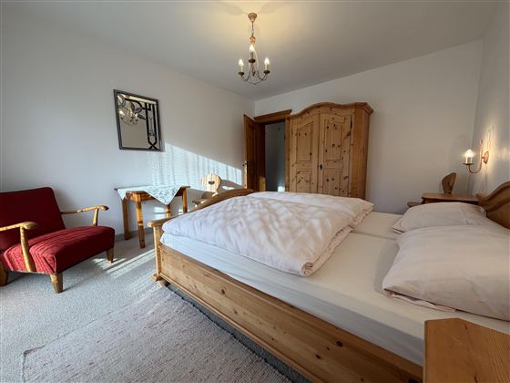 Ein gemütliches Schlafzimmer mit einem großen Bett und einem Holzschrank. Ein roter Sessel und ein kleiner Tisch runden die Einrichtung ab.