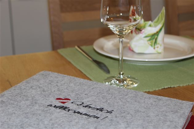 Ein schöner Tisch gedeckt mit einem Glas Wein und einem dekorierten Teller. Auf dem Tisch liegt eine graue Stoffmappe mit dem Schriftzug „Willkommen!“.