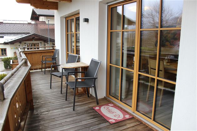 Ein gemütlicher Balkon mit Holzdielen, zwei Stühlen und einem Tisch. Durch die großen Fenster ist ein schöner Blick nach draußen möglich.