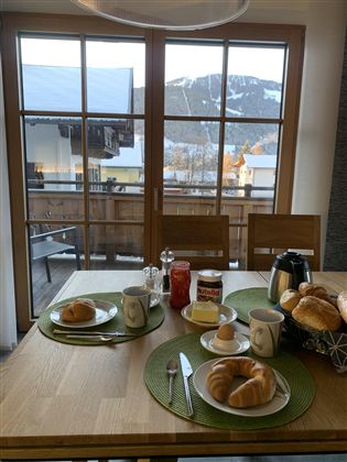 Ein gemütlicher Frühstückstisch mit Brötchen, Marmelade und Getränken. Im Hintergrund sind schneebedeckte Berge und ein Balkon zu sehen.