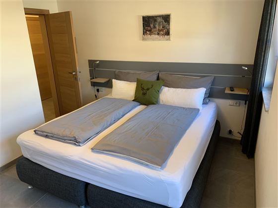 Ein gemütliches Schlafzimmer mit einem großen Doppelbett und modernen Möbeln. An der Wand hängt ein Bild und es gibt Fenster mit Vorhängen.