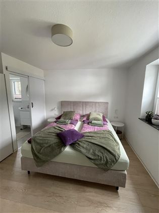 Een moderne slaapkamer met een groot tweepersoonsbed en kleurrijke kussens. De kamer is licht en vriendelijk, met een raam dat veel licht binnenlaat.