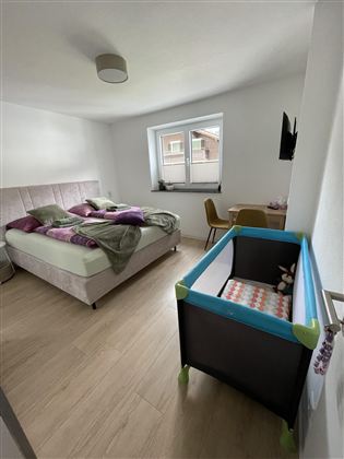 Een moderne slaapkamer met een groot bed en een gezellige deken. In de hoek staat een kinderbed en een klein bureau.