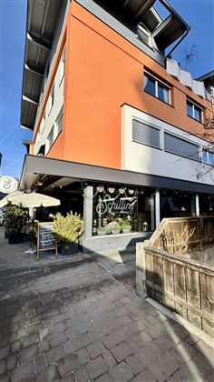 Moderní budova s oranžovou fasádou. Před vchodem stojí stůl se slunečníkem a rostlinami.