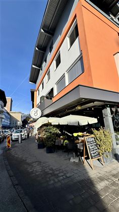 Moderní kavárna na rohu ulice s terasou pod slunečníkem. Fasáda je natřená šedě a oranžově a zve k posezení.