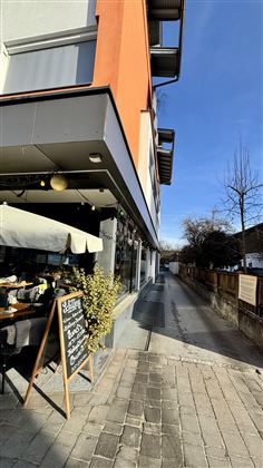 Moderní kavárna s terasou a černou tabulí před vstupními dveřmi. Jasné nebe a klidná ulice dodávají scéně uvolněnou atmosféru.