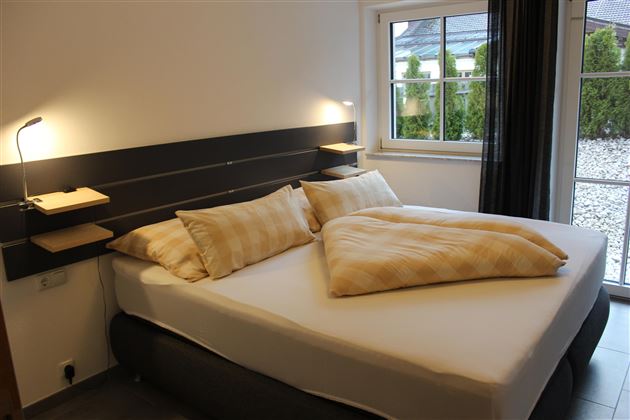 Een moderne slaapkamer met een groot lits-jumeaux en twee kussens. Grote ramen zorgen voor veel daglicht en uitzicht op buiten.