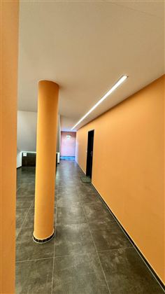 Prostorná chodba s oranžovými zdmi a barevnými sloupy. Podlaha je tmavá a na konci chodby je dveře.