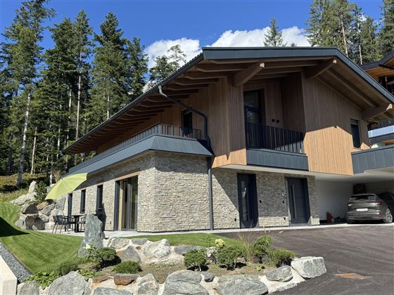 Een modern huis met een houten en stenen gevel. Het ligt in een groene omgeving met bomen en een heldere lucht.